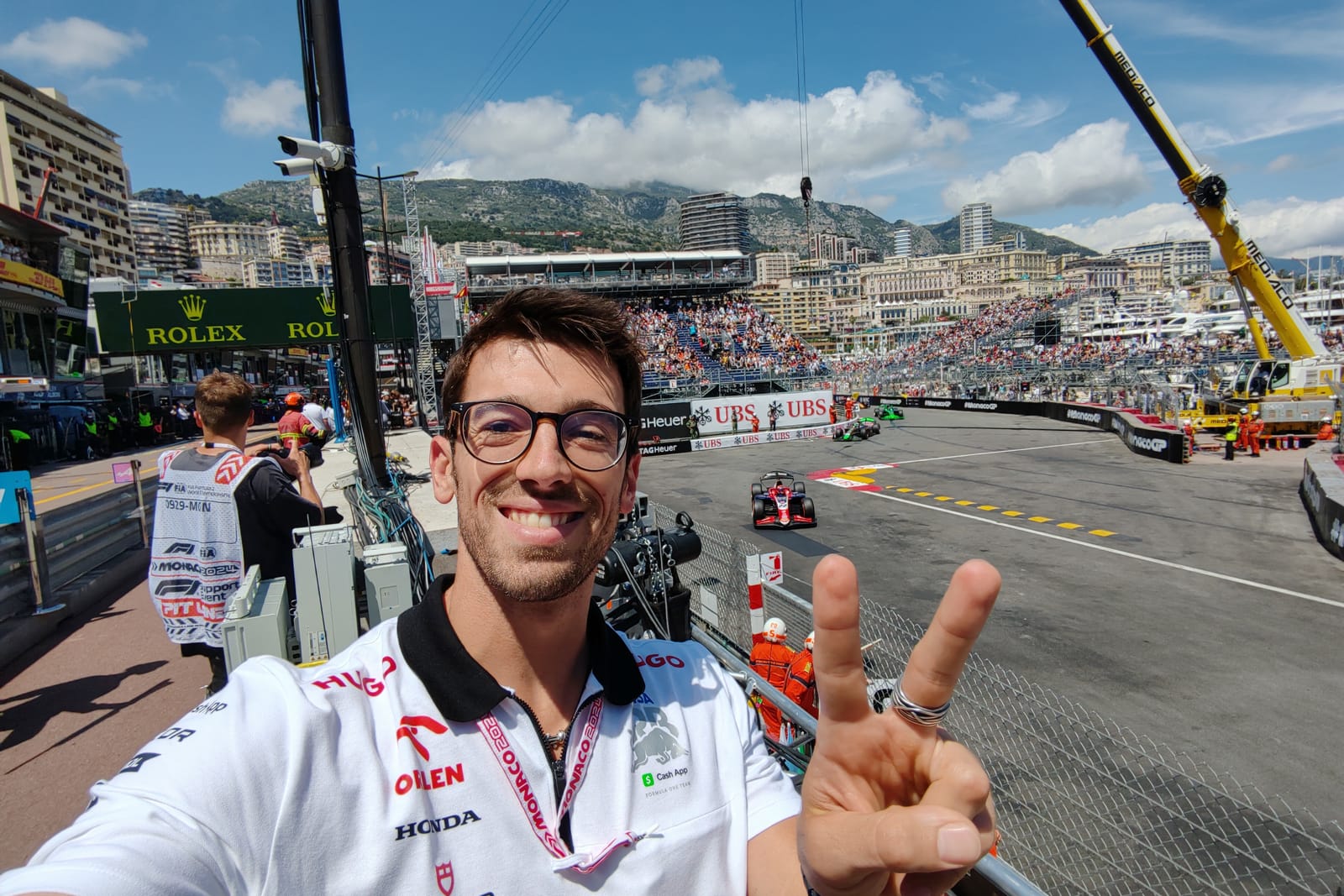 Matteo Tomei nella corsia dei box del Gran Premio di Monaco, con la divisa della squadra; dietro di lui una monoposto in pista, le tribune e i palazzi di Monte Carlo.